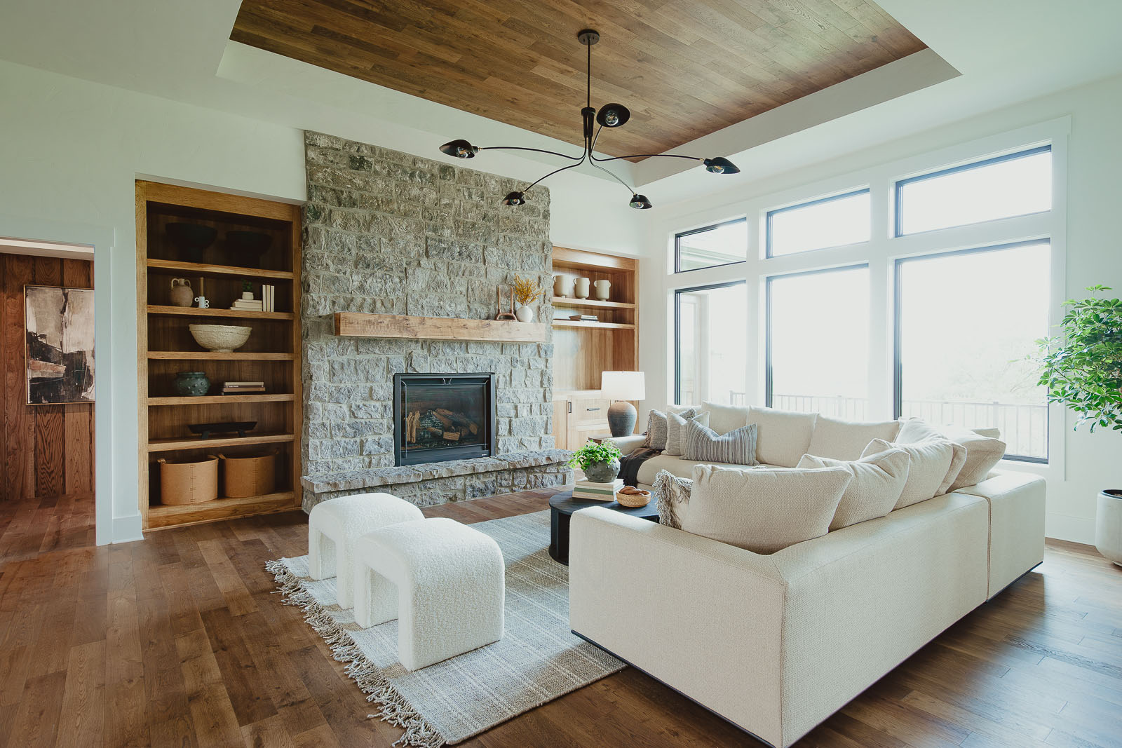 This warm, inviting model home family room has a large stone fireplace, framed by built-in shelves. A large cream colored L-shaped sectional sits in front of the fireplace along with two u-shaped ottomans and a large coffee table. Off the left of the photo you can view the hallway to the primary bedroom.