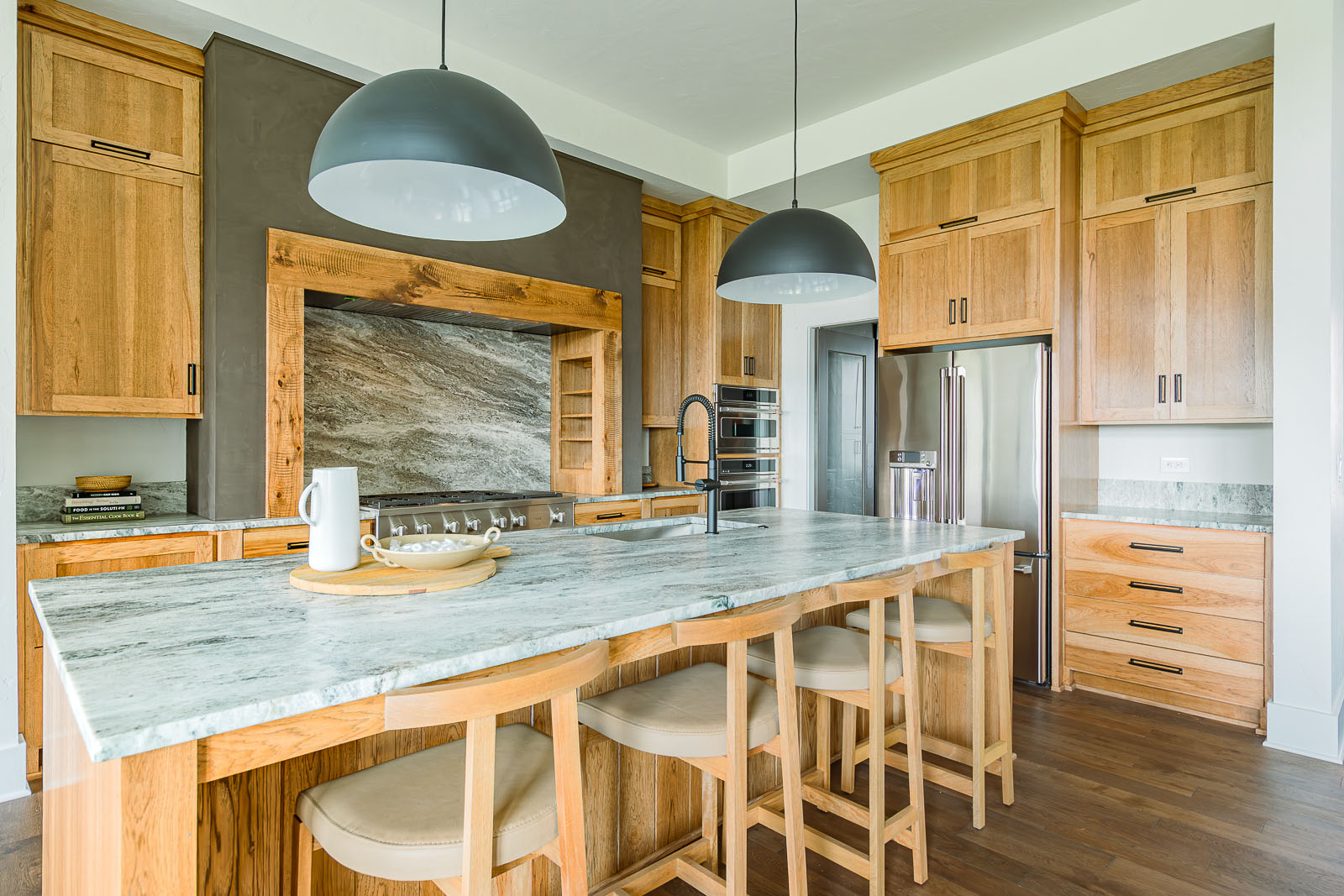 Kitchen with four barstools, custom cabinetry and black bowl-shaped pendant lights. This warm inviting model home was designed by Sara Svehla Interior Design for the 2023 Fall Model Home Tour in North Kansas City, Missouri.
