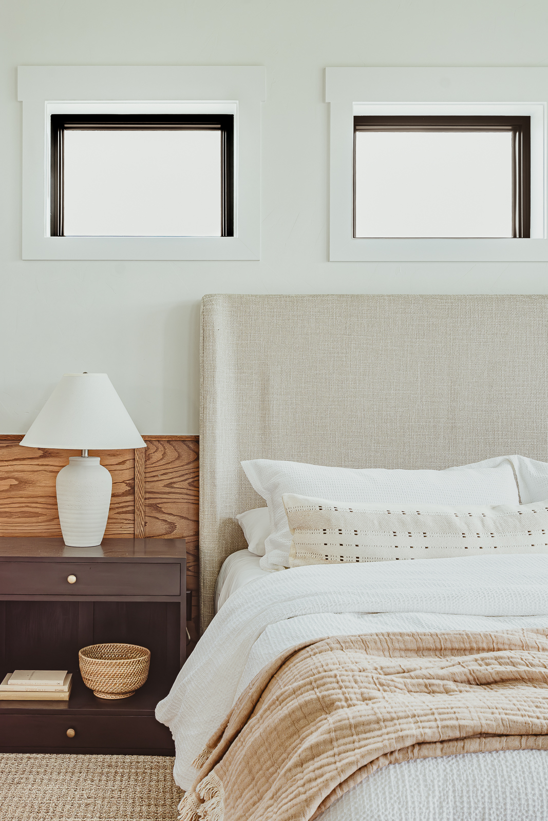 This warm, inviting primary bedroom with wood paneled wainscoting overlooks a golf course in north Kansas City. It was part of the 2023 Fall Model Home Tour and was designed by Sara Svehla Interior Design. The king bed boasts a 5 foot tall, slightly inward curving headboard in cream upholstery. A dark mahogany colored nightstand and a single cream ceramic lamp sit atop it. 