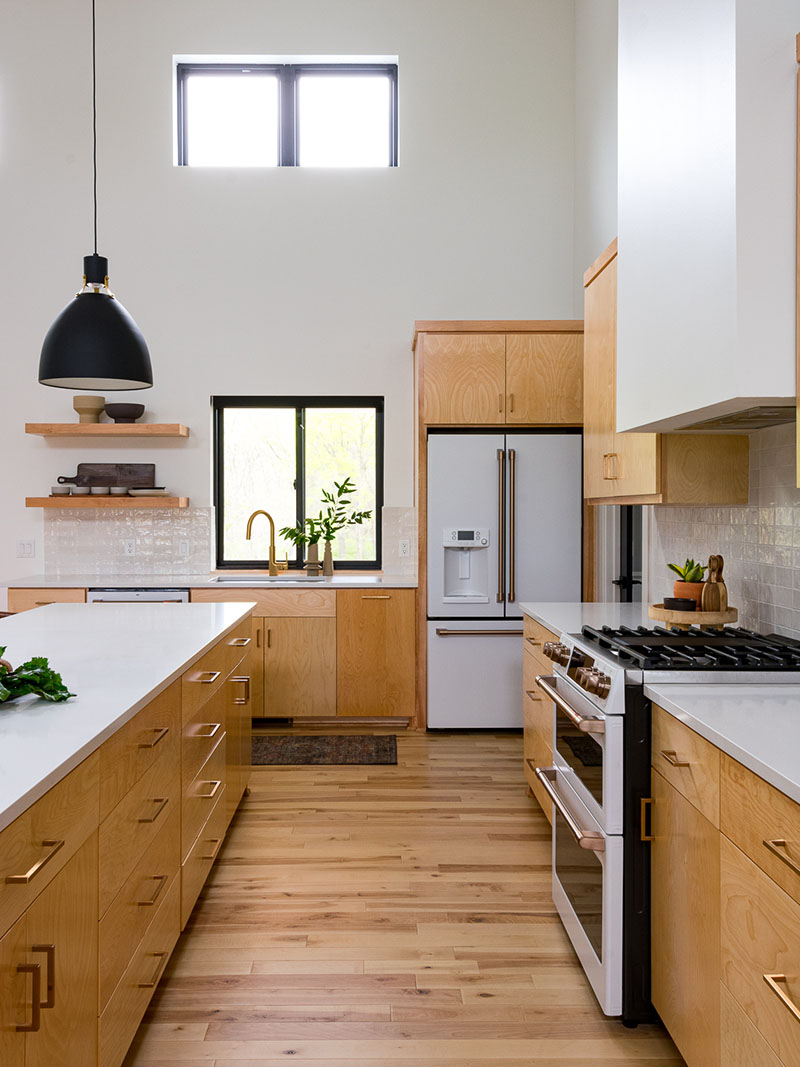 Bright white walls and countertops accented by light colored custom cabinets with brass hardware and matte black pendants over the kitchen island. Floating shelves to the left of the kitchen sink overlooking the back patio in this bright, airy kitchen renovation.