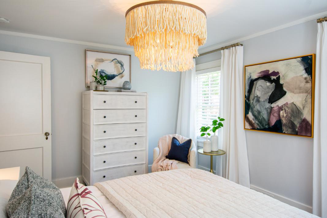 Primary bedroom with pastel color artwork in a gold frame, showing the bottom corner of the king bed with plush bedding and an accent chair in front of a window with custom drapery.