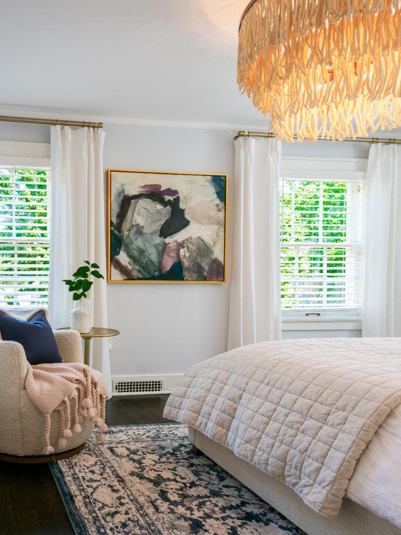 Primary bedroom with pastel color artwork in a gold frame, showing the bottom corner of the king bed with plush bedding and an accent chair in front of a window with custom drapery.