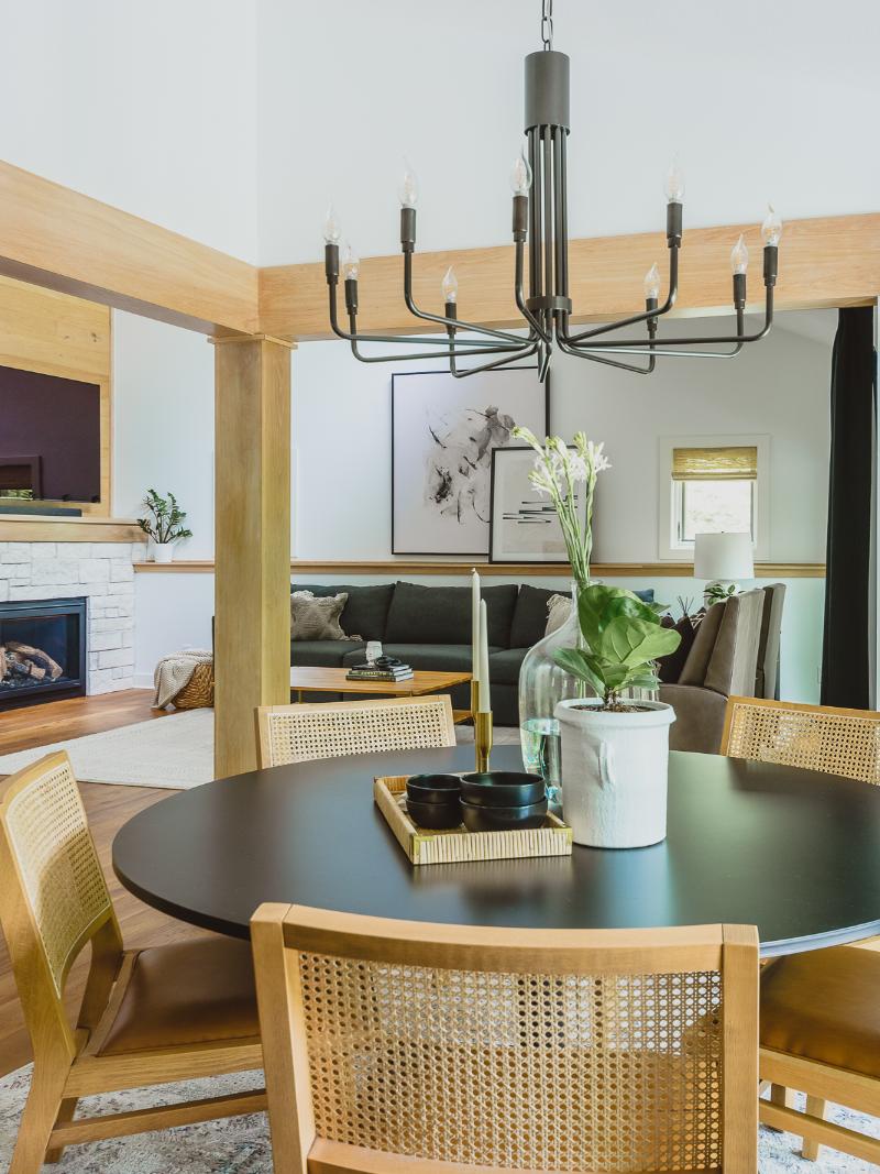 With a neutral palette of soft white walls, oak beams, and natural wicker dining chairs around a small round dining table, overlooking a spacious background in this clean and bold interior.