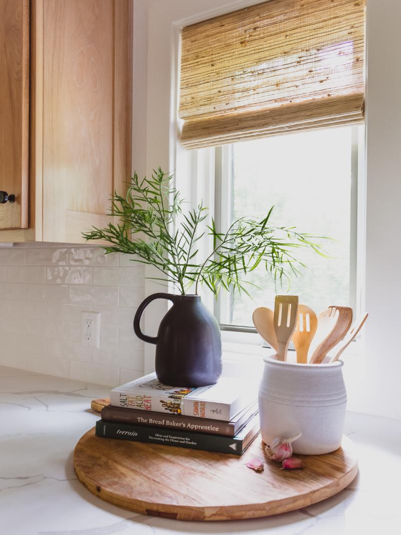 Cookbooks and a vase of greenery sit atop a round cutting board, alongside a crock of wooden utensils in this clean and bold interior designed by Sara Svehla Interior Design.
