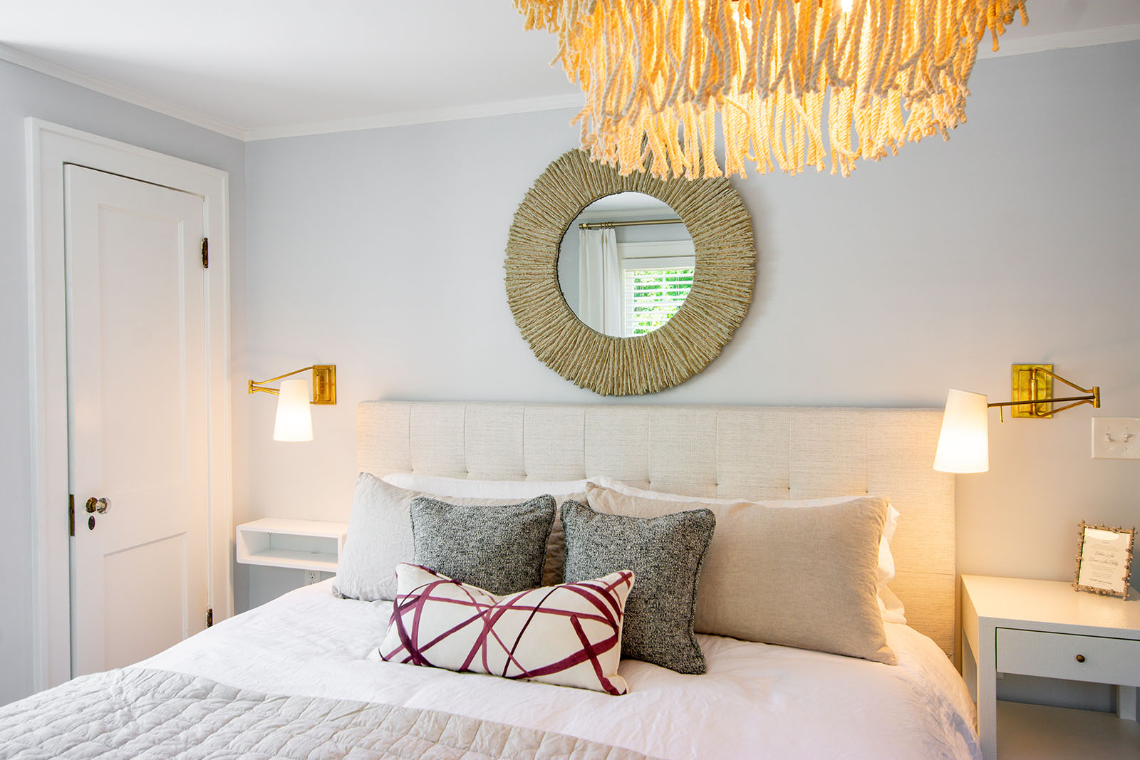 King bed framed by two custom floating shelves for nightstands with a fringe chandelier and a natural weave mirror hanging over the headboard.