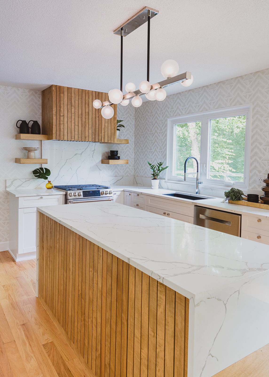 This custom renovated kitchen is featured on the Interior Design for Homeowners page for Sara Svehla Interior Design's website. The image showcases a gorgeous bubble chandelier over a marble-esque waterfall island. The kitchen has lots of custom wood slats and a white herringbone tile on the walls.